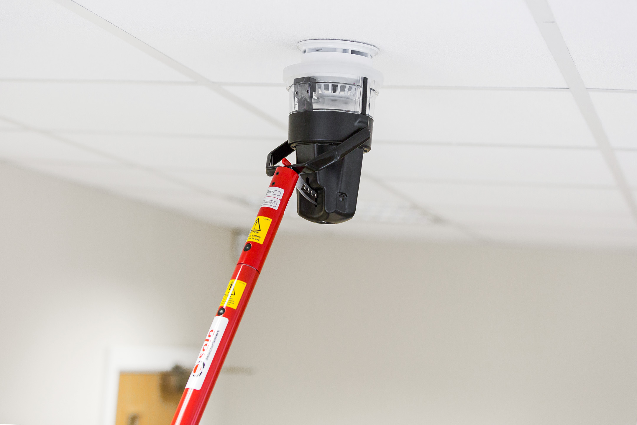 black solo 461 battery powered heat detector tester attached to a red solo telescopic access pole testing a smoke detector