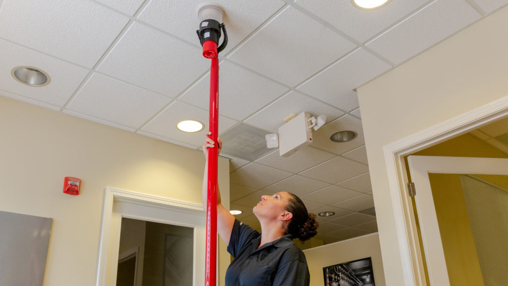female holding a red telescopic access pole that is attached to a solo 330 aerosol dispenser and testing a smoke detector